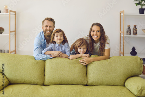 Portrait of happy family at home. Cheerful young European mother, father and children standing together by modern green sofa in living room, looking at camera and smiling