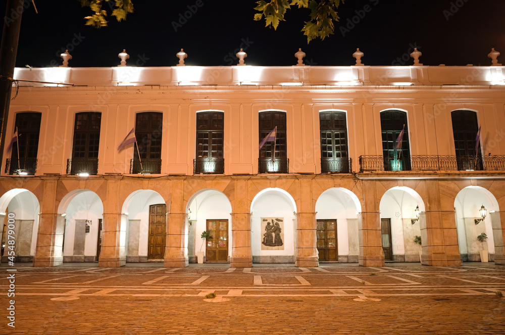 Fototapeta premium Cordoba, Argentina - January, 2020: Part of city hall of Cordoba building called Cabildo de Cordoba at night with lighting and Argentinian flags waving in wind. Old historic building with archway