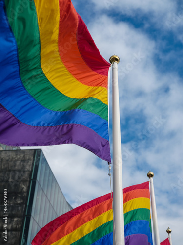 A gay pride flag is blowing in the wind, a blue sky with fluffy clouds in the background.