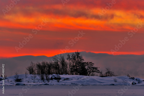 Dramatic scene including fiery sunrise on extremely cold winter morning with sea fog and icy covering Baltic sea behind ice and snow covered island and clouds appearing as if in flame or as ablaze.