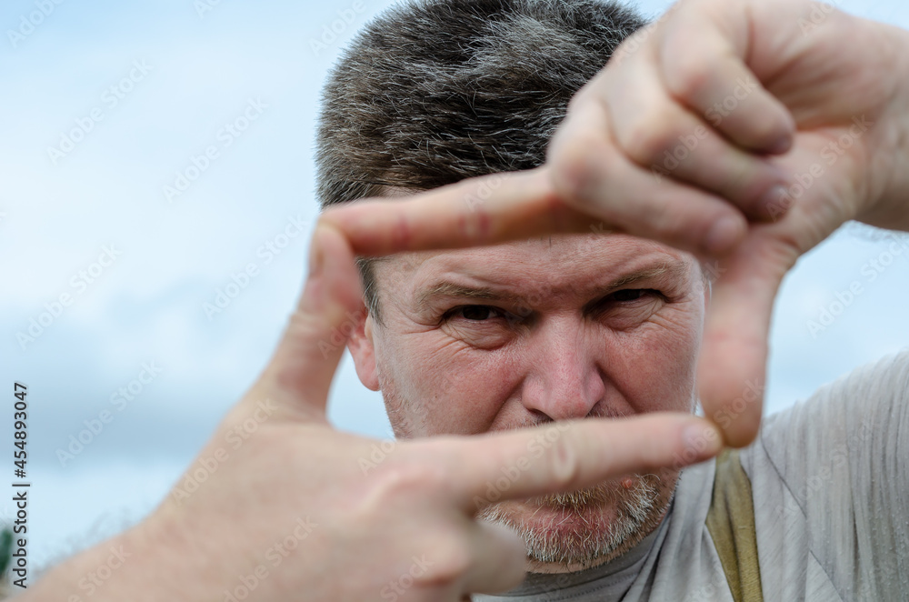Portrait of a grown man making a frame out of his fingers. A gray-haired man looking at the camera through his fingers. Outside the room. Selective focus. Portrait of a grown man making a frame out of his fingers. A gray-haired man looking at the camera through his fingers. Outside the room. Selective focus.