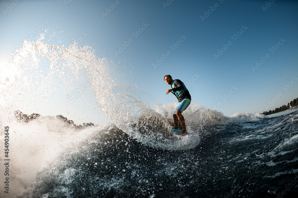 view of the splashing wave along which man rides on wakesurf on the background of blue sky