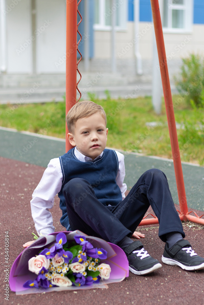 A cute first-grader boy in a school uniform with a beautiful bouquet of colorful flowers in the school yard after the holiday on September 1. Knowledge day. Selective focus A cute first-grader boy in a school uniform with a beautiful bouquet of colorful flowers in the school yard after the holiday on September 1. Knowledge day. Selective focus