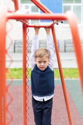 A cute first grader boy at the school on the playground hangs on a horizontal bar on a sunny autumn day. Celebration on September 1st. Knowledge day. Selective focus
