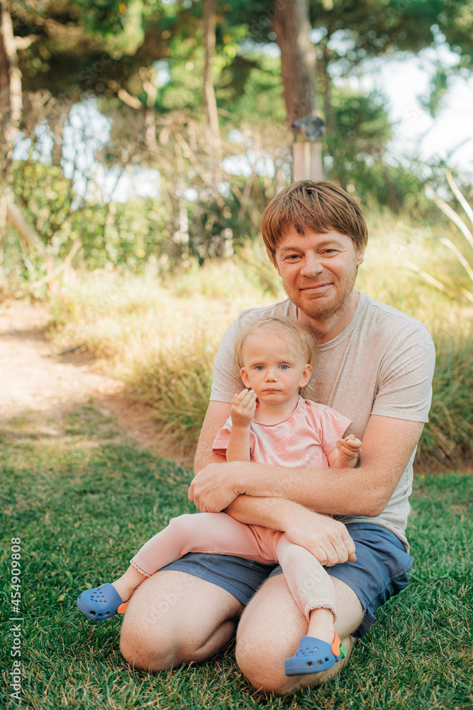 Portrait of happy mid adult man with baby daughter sitting on grass, looking at camera and smiling outdoors. Father walking with his little child. Childhood, family weekend concept Portrait of happy mid adult man with baby daughter sitting on grass, looking at camera and smiling outdoors. Father walking with his little child. Childhood, family weekend concept