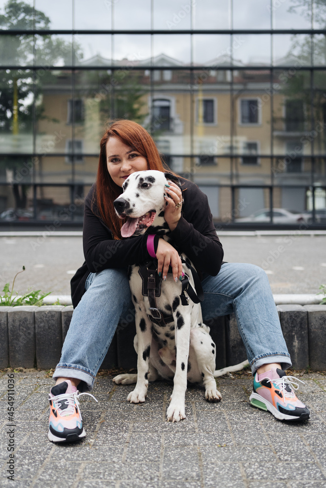 Happy woman sit and cuddling with her dalmatian dog on a glass building background in the city. Friendship, love and care concept