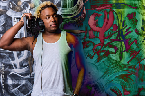 Confident black man in a white T-shirt holds a DJ console in his hands and listens to music with black headphones against the background of graffiti. Professional African-American DJ 