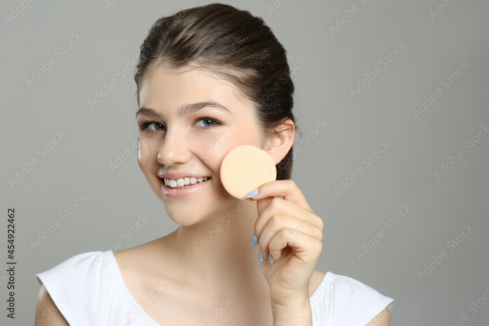 Beautiful girl with foundation smear on her face holding sponge against grey background Beautiful girl with foundation smear on her face holding sponge against grey background