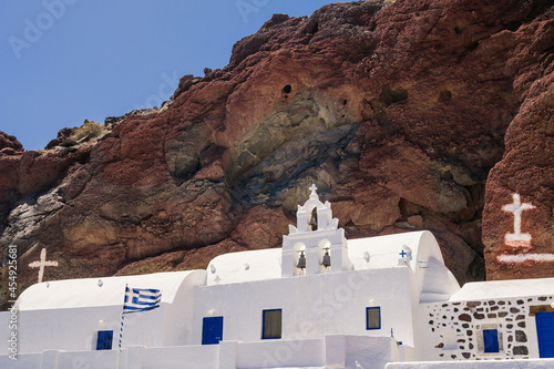 White church with bell tower and blue doors at the foot of red rock. Bottom view. Traditional Orthdox church on Santorini island, Greece. photo