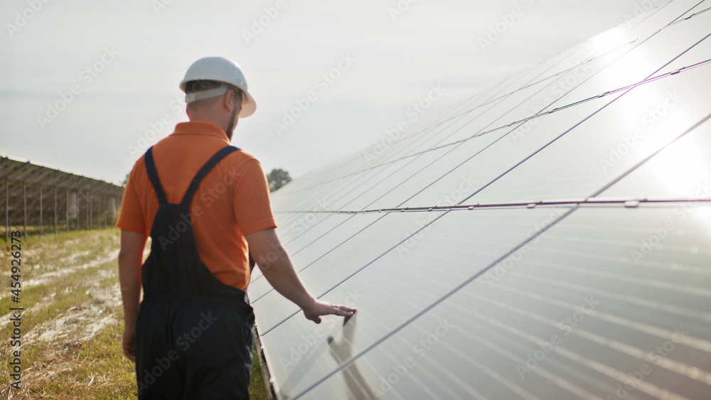 Professional male engineer in protective helmet walk on ecological solar plant construction. Concept of electricity, ecology, technologies. Farm of solar panels. Clean energy production. Green energy Professional male engineer in protective helmet walk on ecological solar plant construction. Concept of electricity, ecology, technologies. Farm of solar panels. Clean energy production. Green energy