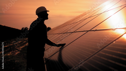 Assistance technical worker in uniform is checking an operation and efficiency performance of photovoltaic solar panels. Unidentified solar power engineer touches solar panels with his hand at sunset