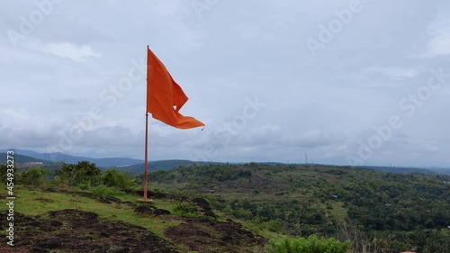 The Hindu religion orange flag tied to the flagpole placed on the top of the hill flies in the strong wind. Shot taken from the famous tourist destination Manjummadikunnu Hill station Kerala India