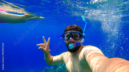 a bearded man in a mask and a breathing tube dives into the red sea against the background of a girl swimming behind him