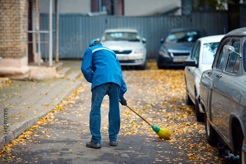 Cleaner removes leafs, janitor with broomstick remove yellow leaves from road. Autumn street sweeper work. Street cleaner sweeping yellow fallen leaves outdoors, autumn seasonal cleaning work photo