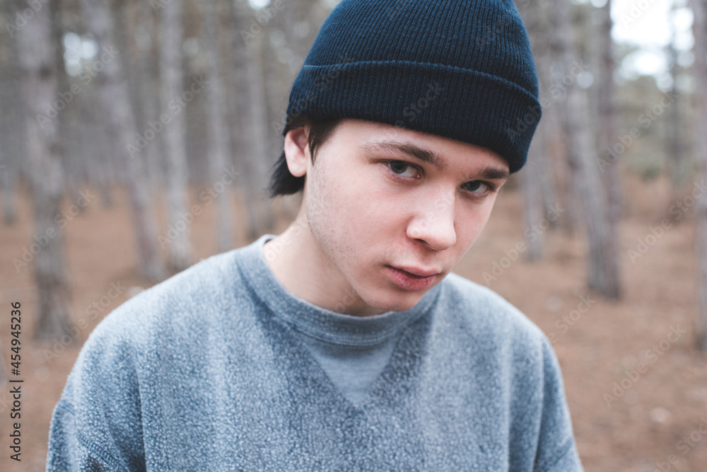 Teenage boy 16-17 year old wear sweatshirt and knit hat posing in woods looking at camera. Teenagerhood. Serious guy posing outdoors. Teenage boy 16-17 year old wear sweatshirt and knit hat posing in woods looking at camera. Teenagerhood. Serious guy posing outdoors.