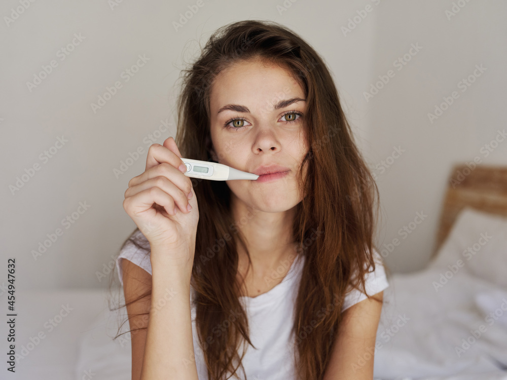 woman holding a thermometer in her mouth while sitting on the bed checking temperature woman holding a thermometer in her mouth while sitting on the bed checking temperature