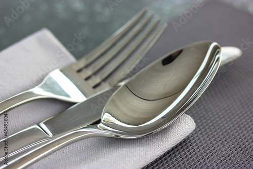 Metal soup spoon, knife, fork lie on a gray napkin on a black grey surface top view. Table setting, cloth in a restaurant, cafe, at home kitchen. Gray textile towel. Flatware close up view from above.