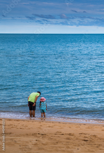 Rear view of father and daughter standing on the beach looking out across the ocean towards a clear blue horizon. 