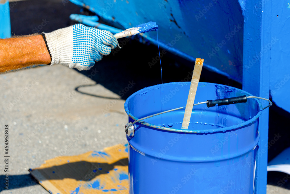Metal bucket with blue paint. A hand in a protective glove holds a paint brush. Painting of metal products. Metal bucket with blue paint. A hand in a protective glove holds a paint brush. Painting of metal products.