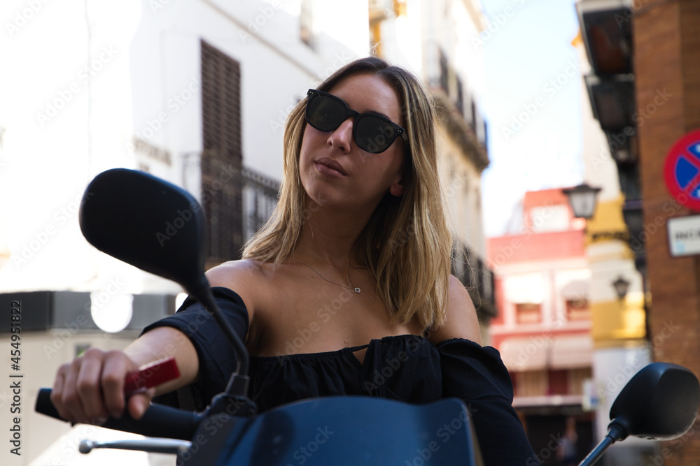 beautiful blonde woman sitting on a red motorbike that she has rented for her holidays in the tourist city. The girl is wearing a black dress and sunglasses. beautiful blonde woman sitting on a red motorbike that she has rented for her holidays in the tourist city. The girl is wearing a black dress and sunglasses.
