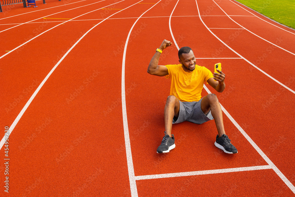 Handsome african american male in yellow t-shirt smiling and raising clenched fist, tensing muscles, taking photo on smartphone. Happy male trainer is sitting at the stadium, take a break from workout