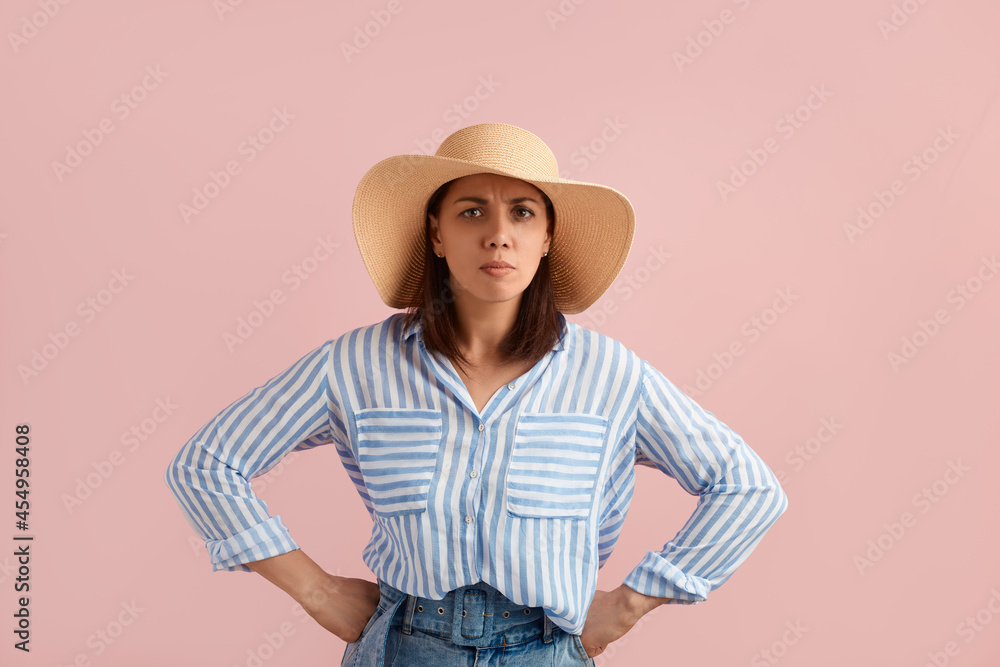 Serious frowning woman brunette keeps hands on waist, stands akimbo, suspects something misleading, wears straw hat, striped shirt, jeans, on pink background. Summer emotions concept. Serious frowning woman brunette keeps hands on waist, stands akimbo, suspects something misleading, wears straw hat, striped shirt, jeans, on pink background. Summer emotions concept.