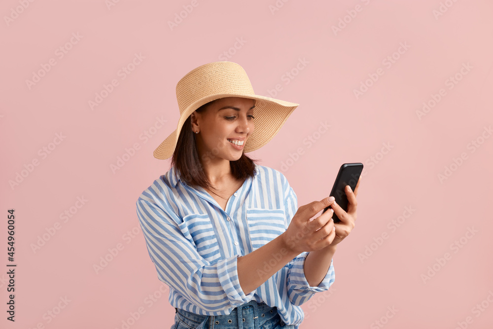 Happy smiling woman is looking at mobile phone, received good news, chatting with her friend, found deal, wears straw hat, striped shirt, jeans with belt, on pink background. Summer emotions concept.