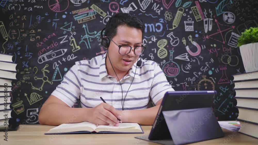 Front view of Asian man wearing glasses having a meeting online on tablet, taking note and using wireless microphone on the table at home, Trying to understand work details. Working at home concept Front view of Asian man wearing glasses having a meeting online on tablet, taking note and using wireless microphone on the table at home, Trying to understand work details. Working at home concept