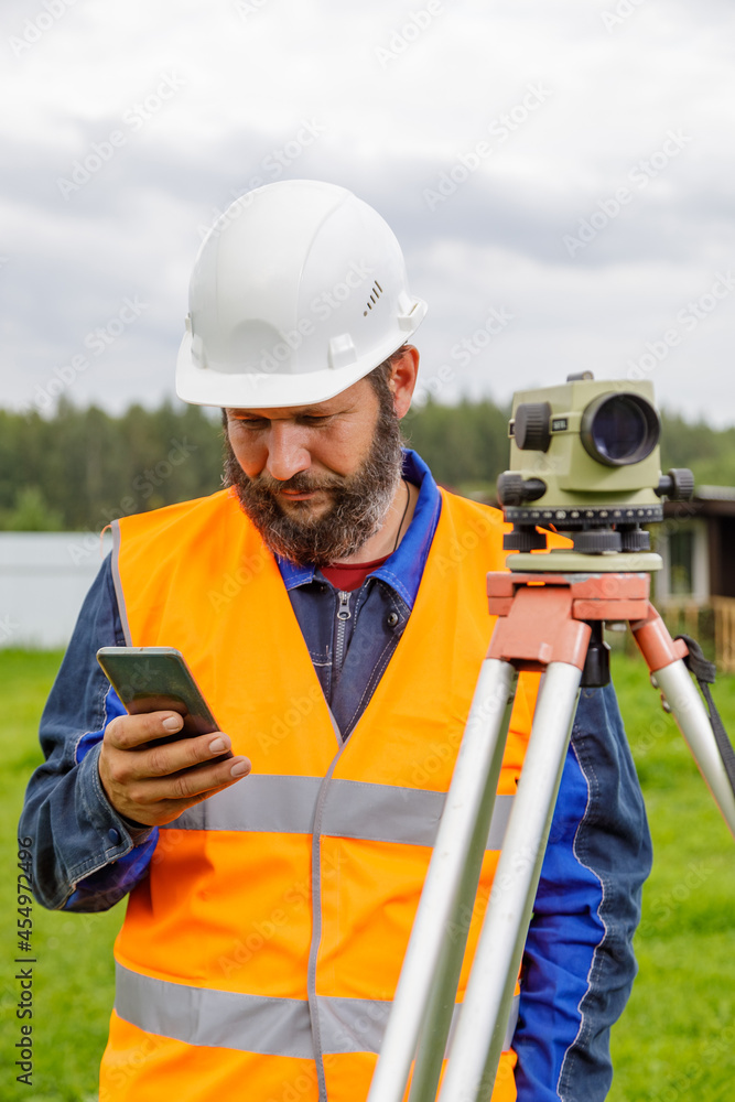 A civil engineer with an optical level looks into a mobile phone. A bearded man is looking for information on his mobile phone.