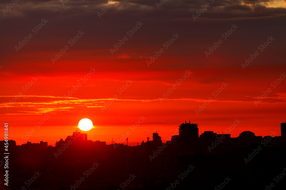 A bright disc of sun rises in the early morning over the city's silhouettes of houses, filling the sky with red light.