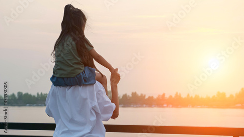 Mommy in loose t-shirt holds toddler daughter on shoulders looking at calm river on city embankment in summer evening twilights backside view