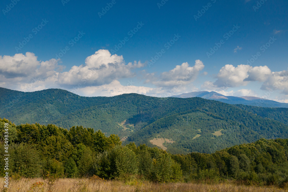 Mountain meadow in the light of the morning sun. Rural summer landscape with a valley in the fog behind the forest on a grassy hill. Fluffy clouds in the bright blue sky. Nature freshness concept. Car