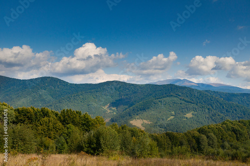 Mountain meadow in the light of the morning sun. Rural summer landscape with a valley in the fog behind the forest on a grassy hill. Fluffy clouds in the bright blue sky. Nature freshness concept. Car © Brylynskyi