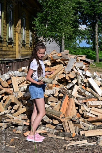 beautiful girl with long hair in white T-shirt and denim shorts puts firewood in pile. Concept of harvesting firewood for winter