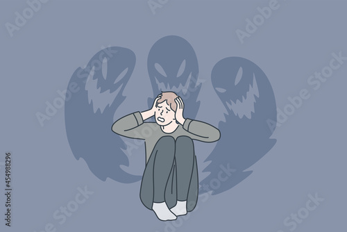 Phobias and inner fears concept. Young stressed man sitting touching head feeling bad with ghosts at wall from inside fears vector illustration 
