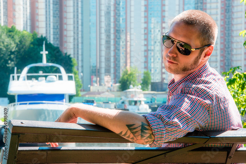 A man with a spider web tattoo in sunglasses sits on a bench in front of a harbor with boats and tall buildings