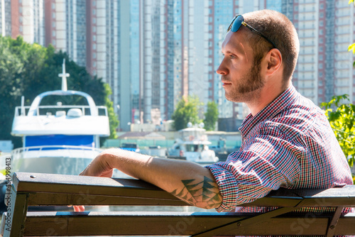 A man with a spider web tattoo in sunglasses sits on a bench in front of a harbor with boats and tall buildings