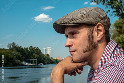 Schilderij op canvas A man in a cap with sideburns is thinking by the river against the background of