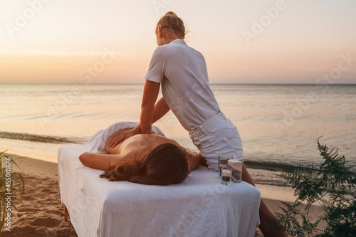 A girl-master of a spa massage at dawn on the seashore makes a back massage to a woman who lies on a massage table. Nearby vases of flowers