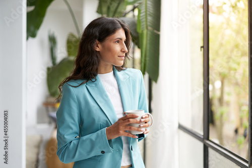 Young business woman satisfied with a job well done relaxing with her morning coffee or tea, looking out the window. Beautiful latina woman celebrating successes on break from work at the office. photo