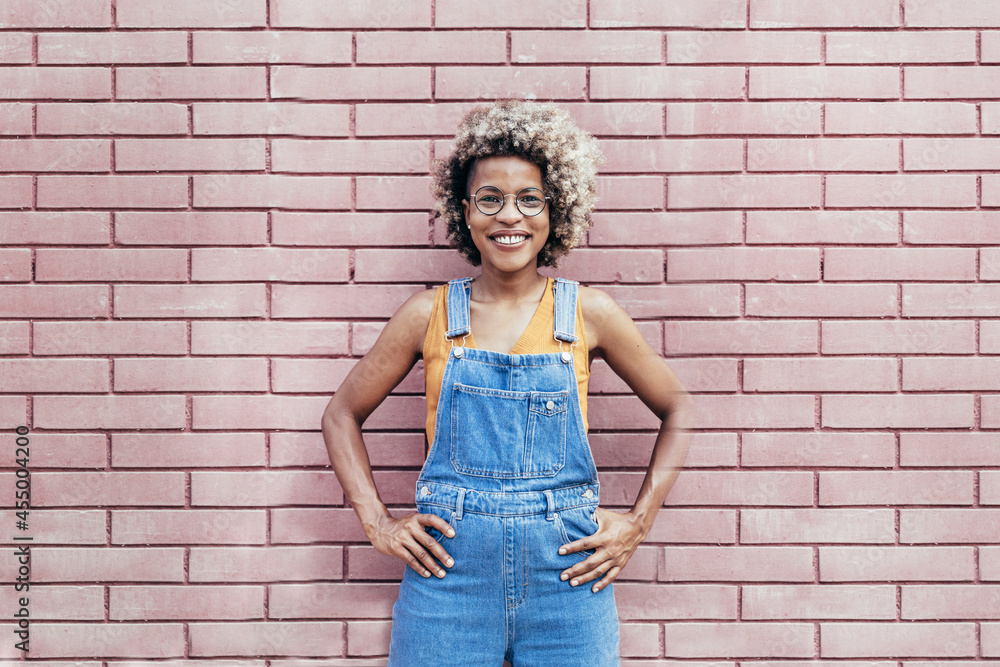 Smiling Black female standing with hands on waists against brick wall outdoors Smiling Black female standing with hands on waists against brick wall outdoors