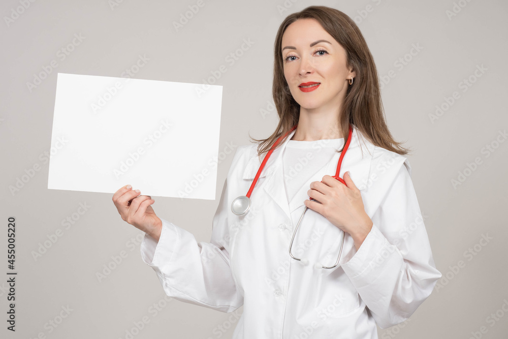 doctor demonstrates sheet of paper. Blank sheet with copy space. Doctor woman on gray background. She smiles and looks into camera. Concept - doctor recommends something. Clinic employee. doctor demonstrates sheet of paper. Blank sheet with copy space. Doctor woman on gray background. She smiles and looks into camera. Concept - doctor recommends something. Clinic employee.