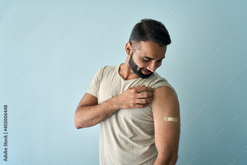 Happy healthy young Indian man showing bandage plaster on arm shoulder after getting vaccination standing on background. Vaccine and people inoculation in India, immunity for covid prevention. Happy healthy young Indian man showing bandage plaster on arm shoulder after getting vaccination standing on background. Vaccine and people inoculation in India, immunity for covid prevention.