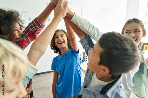 Happy diverse multiethnic kids junior school students group giving high five together in classroom. Excited children celebrating achievements, teamwork, diversity and friendship with highfive concept. © insta_photos