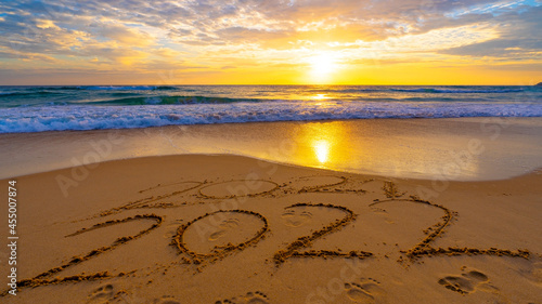 Happy New Year 2022, Lettering on the beach with waves and sunset sky Numbers 2022 year on the seashore, Message hand written in the golden sand on beautiful sunset or sunrise golden sky background.