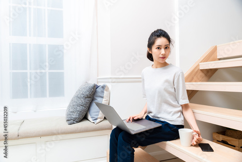 Attractive smiling Asian female sitting on ladder using computer and drinking coffee