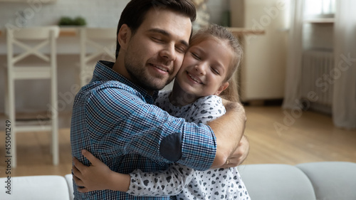 Happy excited father embracing adorable daughter kid with closed eyes. Loving girl hugging beloved daddy on couch with affection, gratitude, tenderness. Fatherhood, childhood, family