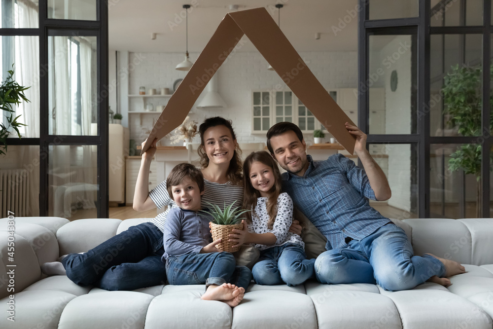 Portrait of happy family couple and two kids making cardboard roof of house above heads, resting, playing on couch, looking and camera, smiling. Parents and children celebrating moving into new house