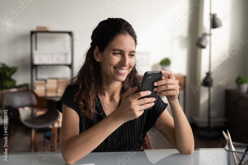 Happy businesswoman using smartphone at office desk, reading message, texting, smiling at screen. Millennial Latin woman chatting, shopping online, making video call. Digital communication