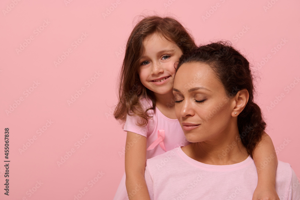 Beautiful baby girl in pink dress with a pink ribbon tenderly hugs her mother with closed eyes, looking at camera, showing support to cancer patients and survivors, pink background with copy space Beautiful baby girl in pink dress with a pink ribbon tenderly hugs her mother with closed eyes, looking at camera, showing support to cancer patients and survivors, pink background with copy space